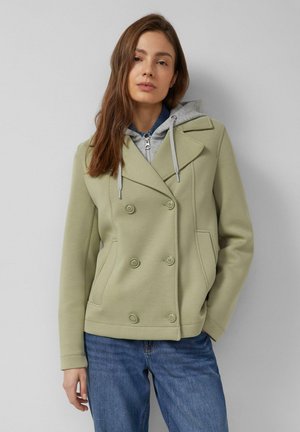 Kobieta o długich brązowych włosach, ubrana w jasnozielony dwurzędowy płaszcz na szarym bluzie z kapturem i niebieskich dżinsach, stojąca na tle szarego tła.