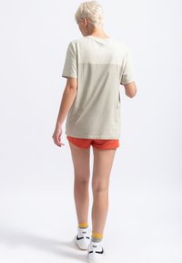 Lichtgrijze t-shirt met een ontspannen pasvorm, voorzien van korte mouwen en een subtiel logo op de achterkant, gecombineerd met oranje shorts en witte sneakers.