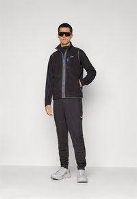 Man in een zwart fleece vest, zwart langarmshirt, zwarte joggerbroeken, witte sneakers en grote zwarte zonnebril, staande tegen een witte achtergrond.