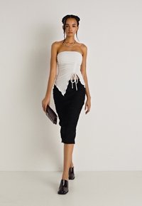 Top alb fără bretele, cu textură plisată și detaliu cu șnur, asortat cu o fustă neagră ajustată, cu un model texturat. Posetă și tocuri maro.