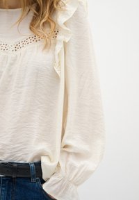 Blusa bianca con scollatura voluminosa e delicati dettagli in pizzo, abbinata a jeans in denim blu e una cintura nera. Tessuto morbido e testurizzato.