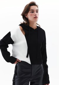 Kapuzenpullover mit einem gesplitteten Design in Schwarz und Weiß, angereichert mit einem strukturierten Strick und einem großen Ausschnitt an der linken Schulter.