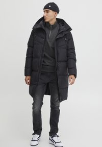 Sort polstret jakke med høj krave, to lynlåslommer, båret over en mørk sweater. Sættet består også af mørke jeans og hvide sneakers.
