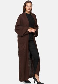 Brun stickad lång kofta med vida ärmar och en framficka, i kombination med en svart crop top och slim fit svarta byxor. Elegant textur och passform.