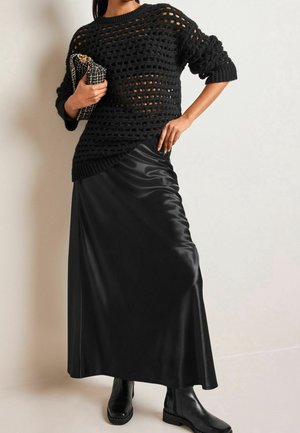 Femme portant un pull noir à maille ouverte, une longue jupe en satin noire, des bottines noires, tenant une pochette chaîne noire et dorée.