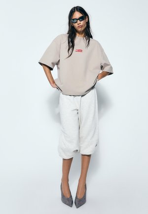 Ženska nosi preveliko bež srajco, bele kratke trenirke, sive visoke pete in črna sončna očala, stoji pred enostavnim belim ozadjem.