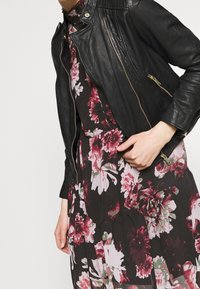 Veste en cuir noire avec des poches zippées et une coupe ajustée, portée par-dessus une robe à motifs floraux avec de grandes fleurs roses et bordeaux.
