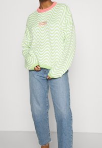 Zielony i biały sweter w faliste wzory z różowym prążkowanym dekoltem, z haftem "RAGGED" w różowym na piersi. Luźny fason i dziana tekstura.