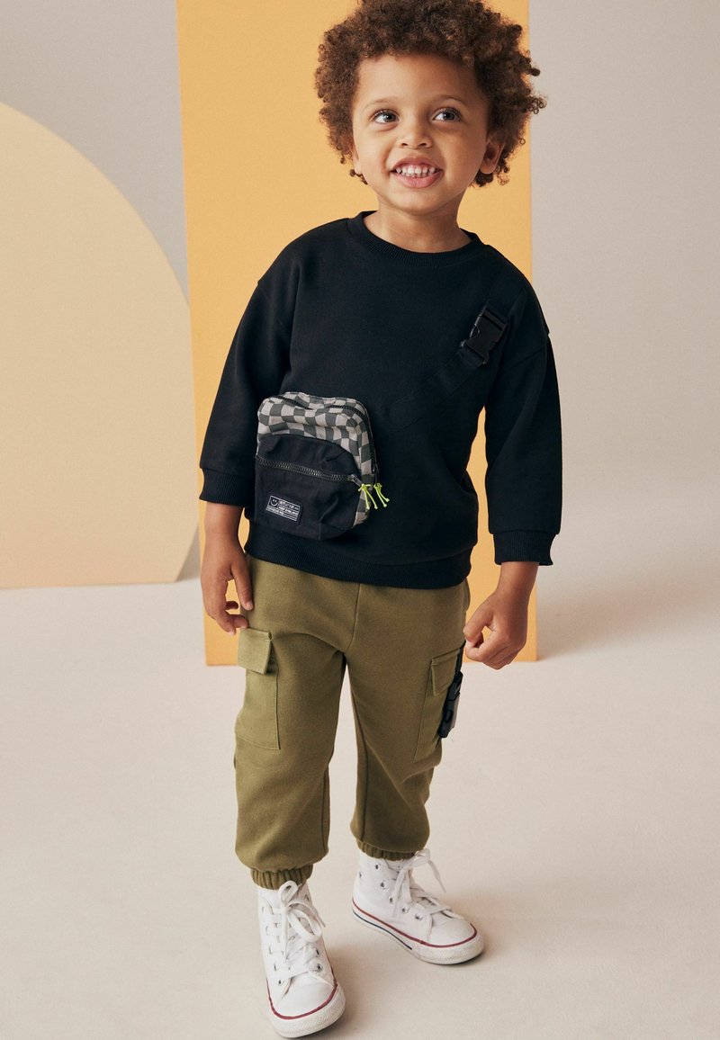Svart sweatshirt med sidoficka, olivgröna cargobyxor med elastiska muddar, och vita högskor. Lockigt hår, leende uttryck.