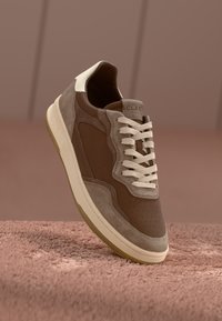 Brown suede sneaker with cream accents, perforated detailing, and white laces. Thick gum sole. Brand name "CLAE" visible on heel.
