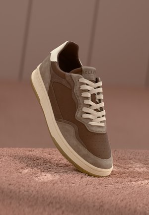 Brown suede sneaker with cream accents, perforated detailing, and white laces. Thick gum sole. Brand name "CLAE" visible on heel.