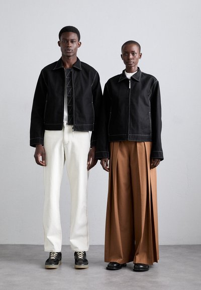 Schwarze Jacken mit kontrastierenden Nähten, weiße Hosen und braune weit geschnittene Hosen. Strukturierte Materialien und unterschiedliche Silhouetten.