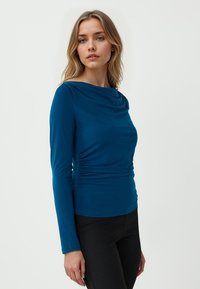 Maglia lunga a maniche con collo a caramella in tonalità teal, con dettagli arricciati sui lati, realizzata in un tessuto morbido, abbinata a pantaloni neri slim-fit.