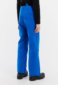 Modré vodoodpudivé lyžiarske nohavice s nastaviteľnými popruhmi, elastickým pásom, viacerými vreckami a voľným strihom, skombinované s čiernymi topánkami.