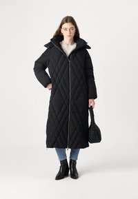 Žena u dugom crnom kviltanom kaputu, plavim farmerkama, crnim čizmama do gležnja, drži crnu pletenu torbu, stoji uz jednostavnu bijelu pozadinu.