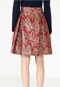Gonna rossa con motivo a foglia d'oro, design plissettato, lunghezza al ginocchio, tessuto strutturato. Indossata con una maglia blu navy, che mostra la vista posteriore.