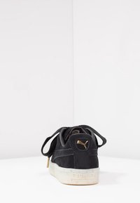 Basket en daim noir à finition texturée, avec un logo doré, des lacets ronds, et une semelle en caoutchouc blanche parsemée de petits éclats.