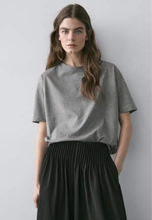 Frau mit langen braunen Haaren, die ein lockeres graues T-Shirt trägt, das in einen hoch taillierten schwarzen Faltenrock gesteckt ist, stehend vor einem schlichten Hintergrund.