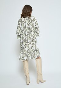 Weißes, floral bedrucktes Kleid mit grünen und braunen Blattmustern, ausgestattet mit einem gestuften Saum. Kombiniert mit kniehohen beigen Stiefeln.