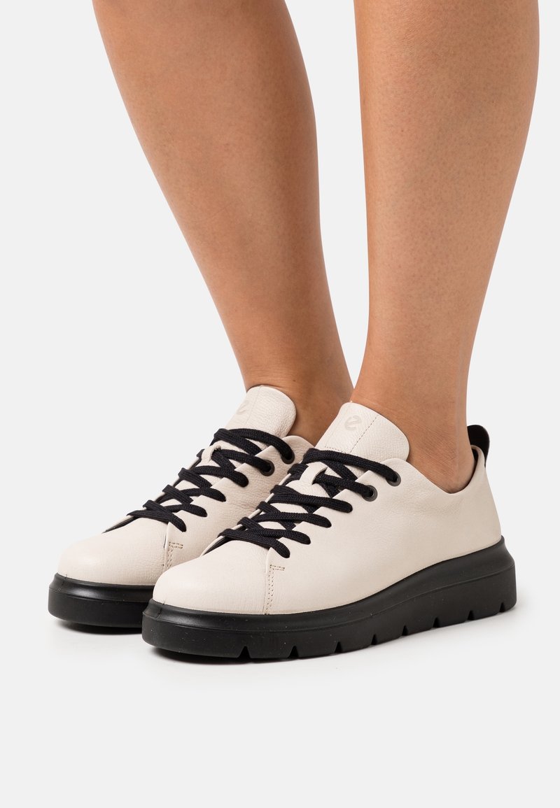 Baskets basses blanches avec lacets noirs et semelles épaisses noires portées pieds nus sur un fond uni.