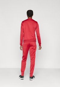 Crveni trenirka od glatke tkanine, s bijelim prugama duž rukava i nogavica, s krojem koji prati liniju tijela i rebrastim manžetama.