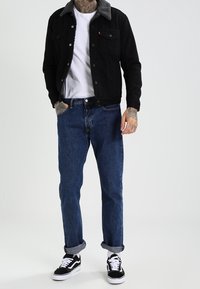 Giacca in denim nero con colletto in pile grigio, T-shirt bianca, jeans blu con risvolto e sneakers bianche e nere. Outfit casual con texture a contrasto.