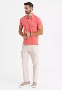 Miestenská ružová polo košeľa s golierom, skombinovaná s béžovými cargo nohavicami s bočnými vreckami a svetlohnedými teniskami. Hladká textúra látky.