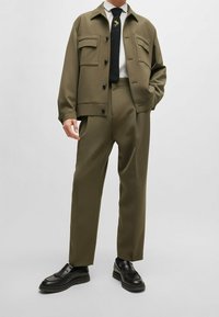 Olijfgroene jas met twee borstzakken en een zwarte das met een bloemenpatroon, gecombineerd met bijpassende olijfgroene broek en zwarte loafers.