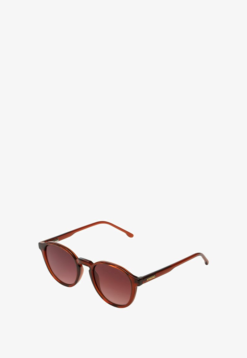 Ronde, bruine, translucente zonnebril met een gradient donker glas en dunne pootjes, met een klein goudkleurig KOMONO-logo op de slaap.