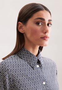 Camicia blu navy e crema con colletto a punta e bottoni bianchi, decorata con forme geometriche su un tessuto texture.