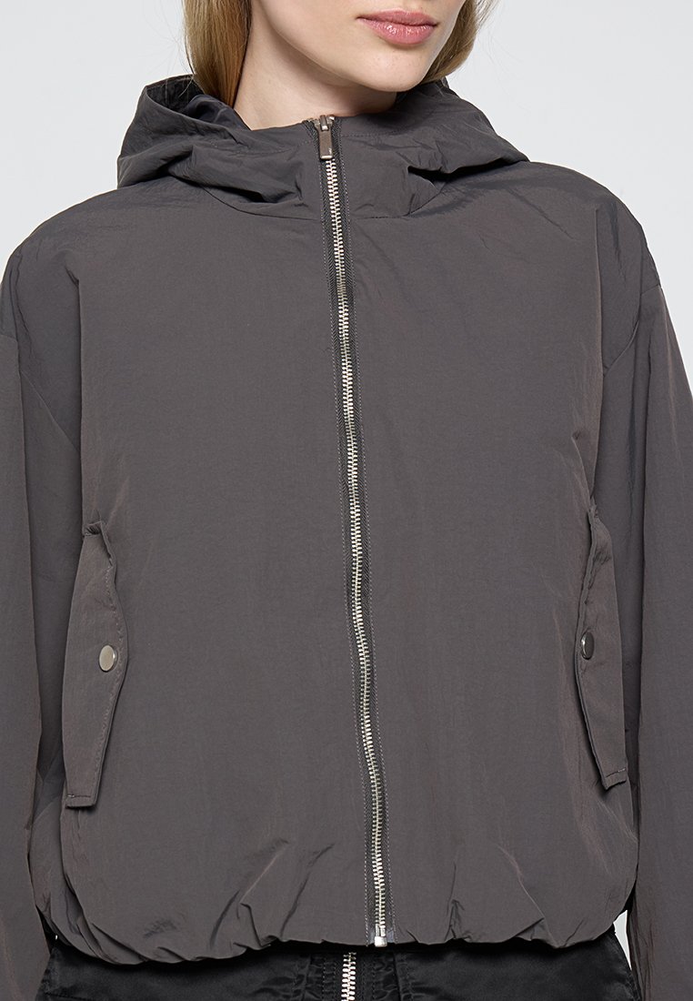 Giacca leggera con cappuccio di colore grigio scuro, dotata di zip completa, tasche con bottoni a pressione e orlo elasticizzato. Realizzata in tessuto liscio e opaco.