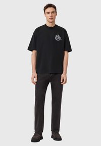 Μαύρο βαμβακερό μπλουζάκι με στρογγυλή λαιμόκοψη και κοντά μανίκια, με λευκό γραφικό δράκο στο αριστερό μέρος του στήθους. Συνδυάζεται με σκούρο γκρι παντελόνια.