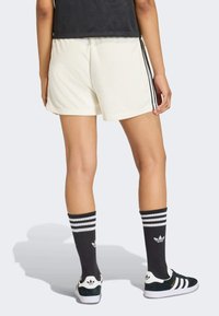 Krémové krátké sportovní šortky s černými bočními pruhy, v kombinaci s černými ponožkami s bílými pruhy a logem Adidas, nošené s černými teniskami.