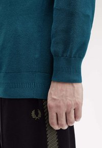 Maglione texturizzato verde acqua con polsini a costine. Abbinato a pantaloni neri con emblema verde e accenti a zigzag lungo il lato.