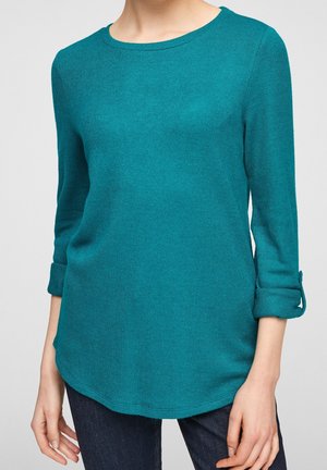 Pull tricoté bleu sarcelle avec des manches trois-quarts retroussées, porté avec un jean foncé, visible du cou aux hanches, visage non visible.