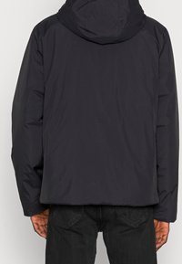 Tmavomodrá prošívaná bunda s pevnou kapucí, která má texturovaný povrch a elastické manžety. Pohled zezadu ukazuje rovný střih bez viditelných log.