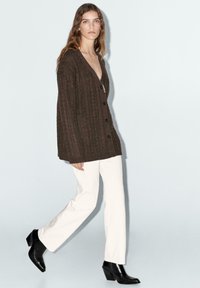 Bruine gebreide cardigan met V-hals en knoopsluiting, gecombineerd met witte rechte broek en zwarte haklaarzen. Minimalistische achtergrond.