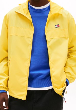 Homme portant une veste jaune zippée par-dessus un pull bleu avec un col blanc, les mains dans les poches de la veste.