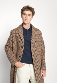 Brun og krem houndstooth frakk med store revers, tre knapper og en avslappet passform, båret over en marineblå polo skjorte og beige bukser.