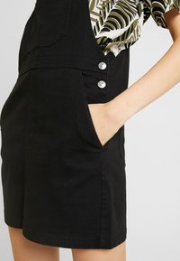 Schwarze Overalls aus Baumwolle mit einer Tasche und zwei silbernen Knopfakzenten. Das Design zeichnet sich durch einen geraden Schnitt und eine strukturierte Oberfläche aus.