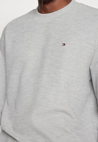 Harmaa huppari, jossa on teksturoitu kangas. Siinä on ribbetty pyöreä kaula-aukko ja pieni logo punaisena, valkoisena ja sinisenä vasemmassa rinnassa.