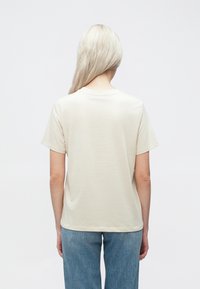 Krems krāsu īsām piedurknēm kokvilnas T-krekls ar apaļu kakla izgriezumu, kuram ir gluda tekstūra un brīva piegriezuma forma, saskaņots ar gaiši ziliem džinsiem.