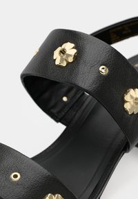 Black leather sandals featuring wide straps adorned with gold floral accents and circular studs, with a flat, textured sole.
