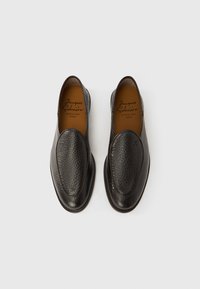 Μαύρα δερμάτινα παπούτσια χωρίς κορδόνια με υφασμένη επάνω επιφάνεια, στρογγυλεμένη μύτη και λείο τακούνι. Το εσωτερικό διαθέτει ανάγλυφο το όνομα της μάρκας και την τοποθεσία.