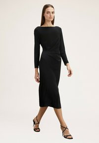 Schwarzes, langärmeliges Kleid mit einem Bootsausschnitt und einem Twist-Detail an der Taille, figurbetonte Silhouette und fließender Stoff, kombiniert mit Riemchenschuhen.