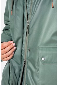 Grön ytterjacka tillverkad av ett slätt, texturerat tyg med ett centralt blixtlås, sidoficka med tryckknapp och justerbar dragsko i huvan.