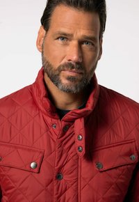 Rote gesteppte Jacke mit hohem Kragen, Druckknöpfen und einer Brusttasche mit silbernen Hardware-Akzenten, aus leichtem Material gefertigt.