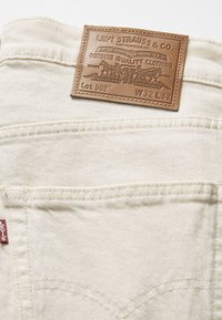 Jeans Levi's 502 en denim blanco roto con un parche de cuero marrón. Presenta un diseño clásico de cinco bolsillos y detalles de costura sutiles.