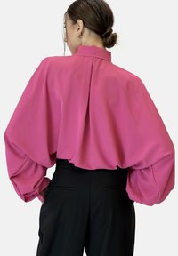Blusa rosa brillante con mangas abullonadas voluminosas y cuello mandarín; cuenta con un profundo pliegue en la parte posterior y una tela suave y lisa. Combinada con pantalones negros.