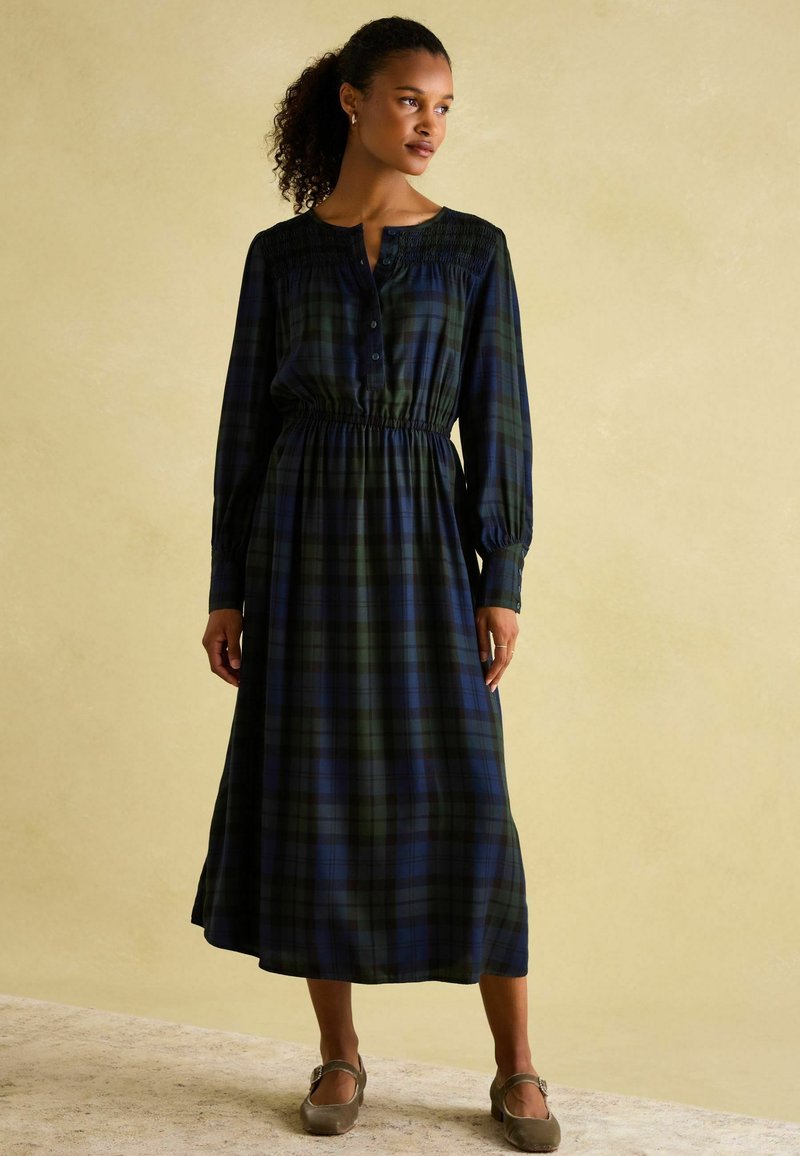 Langes Karo-Kleid mit einem dunkelgrünen und marineblauen Muster, gesmocktem Oberteil, Knopfleiste und langen Ärmeln. Kombiniert mit grauen Riemchen-Schuhen.